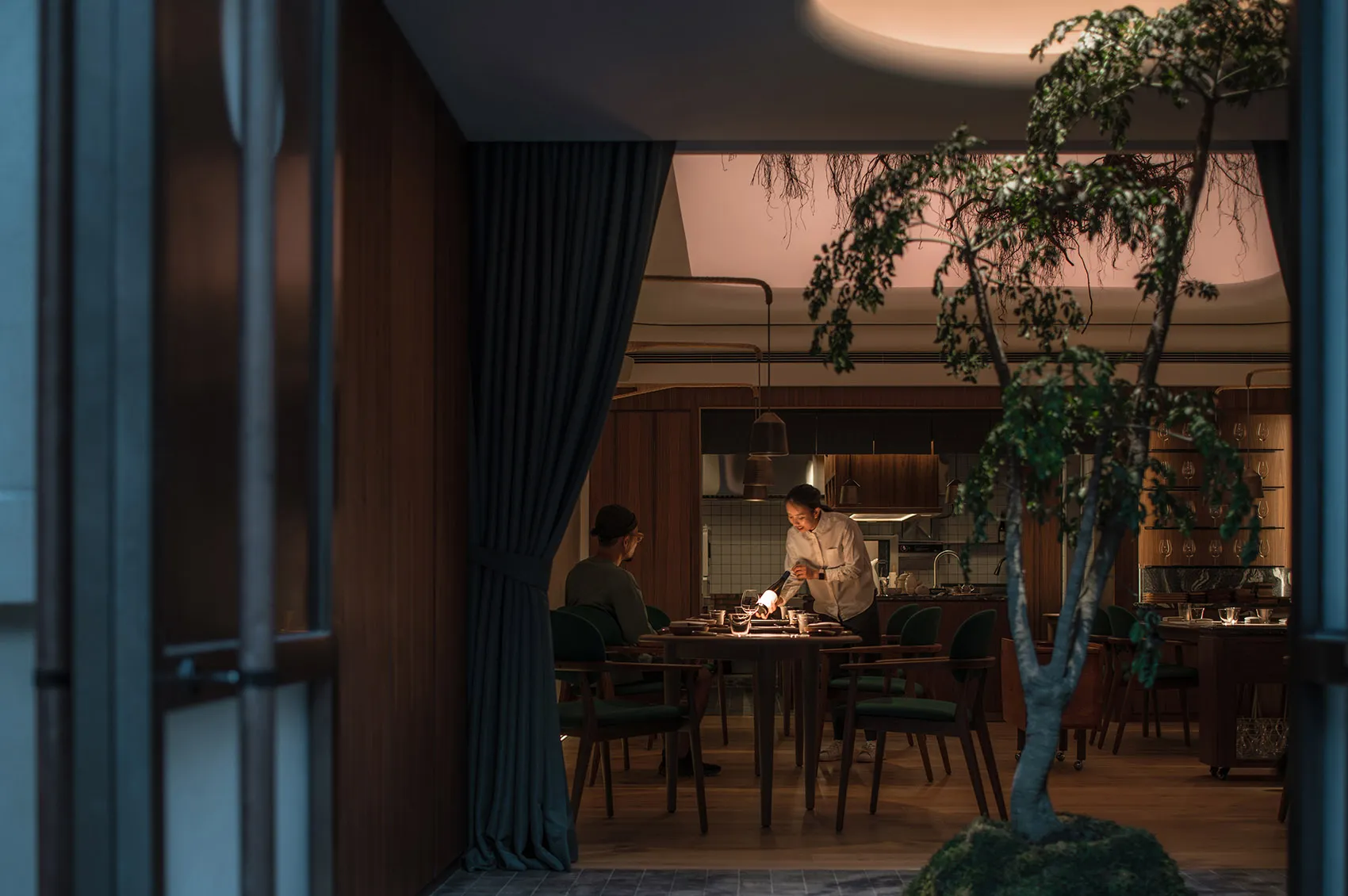 A dark restaurant interior with wooden tables and chairs, greenery, and warm lighting.