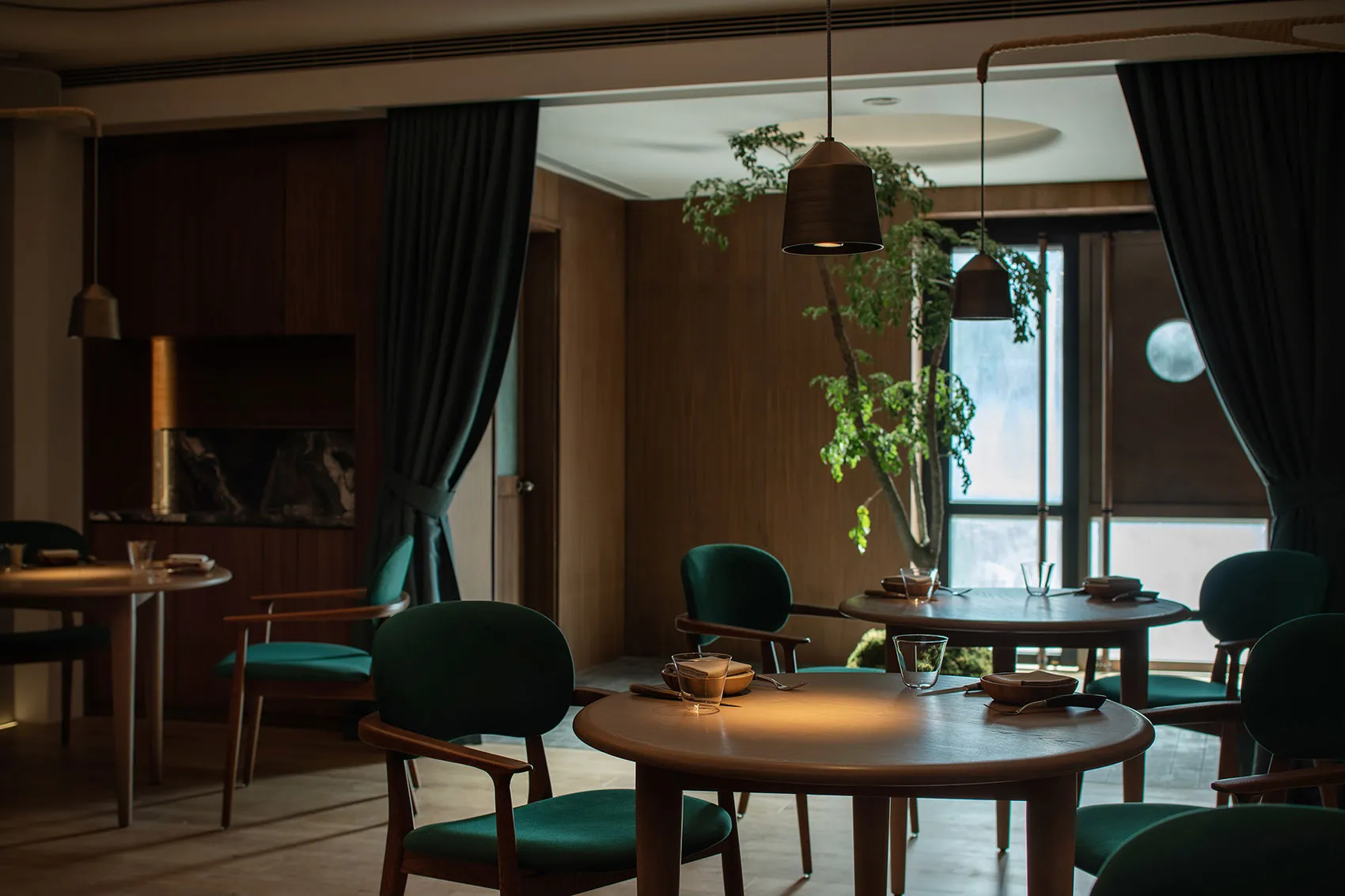 A modern restaurant interior featuring wooden tables, green fabric chairs, and glass accents.