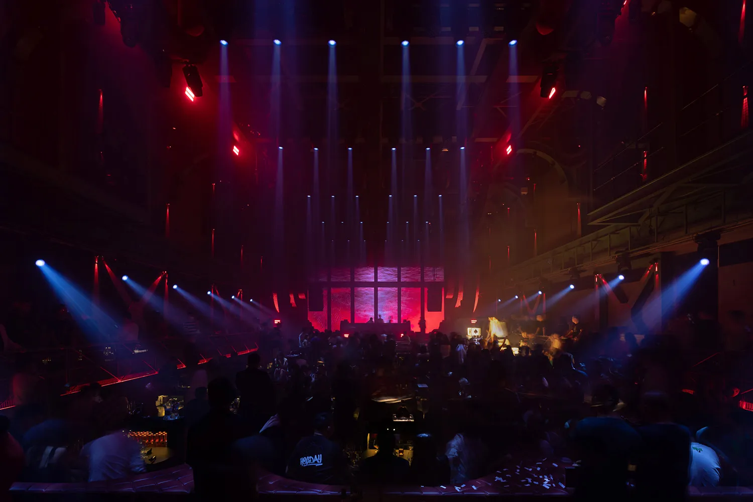A bustling nightclub interior with a cocktail bar illuminated by colorful lights.