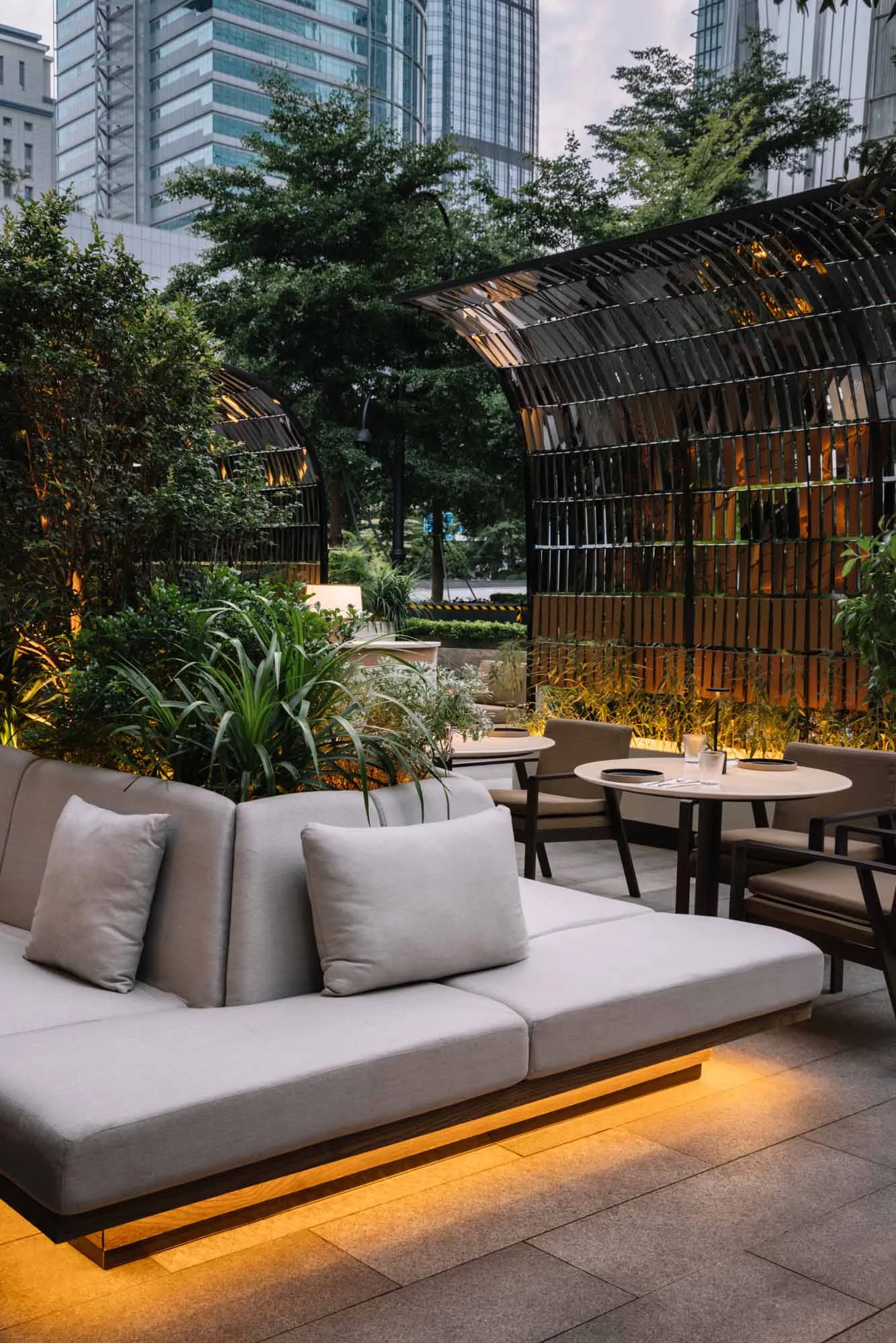 Urban outdoor lounge area with wooden pergola and concrete sofa, featuring greenery and modern seating.