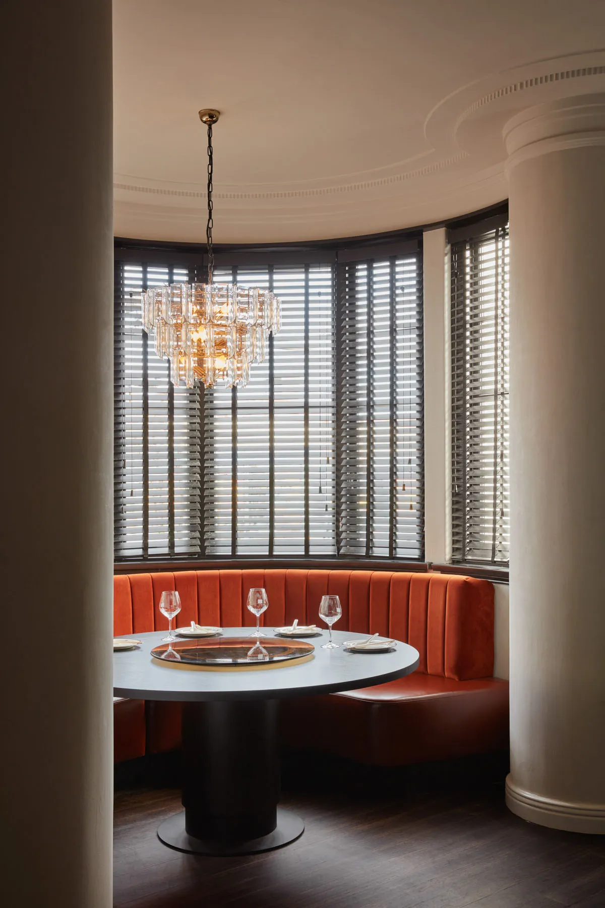 A round dining table with a lazy Susan, glasses, and plates, surrounded by a cozy orange velvet booth seating area with large