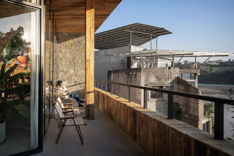 A modern balcony featuring glass railings and wooden structures with a view of brick buildings and natural surroundings.