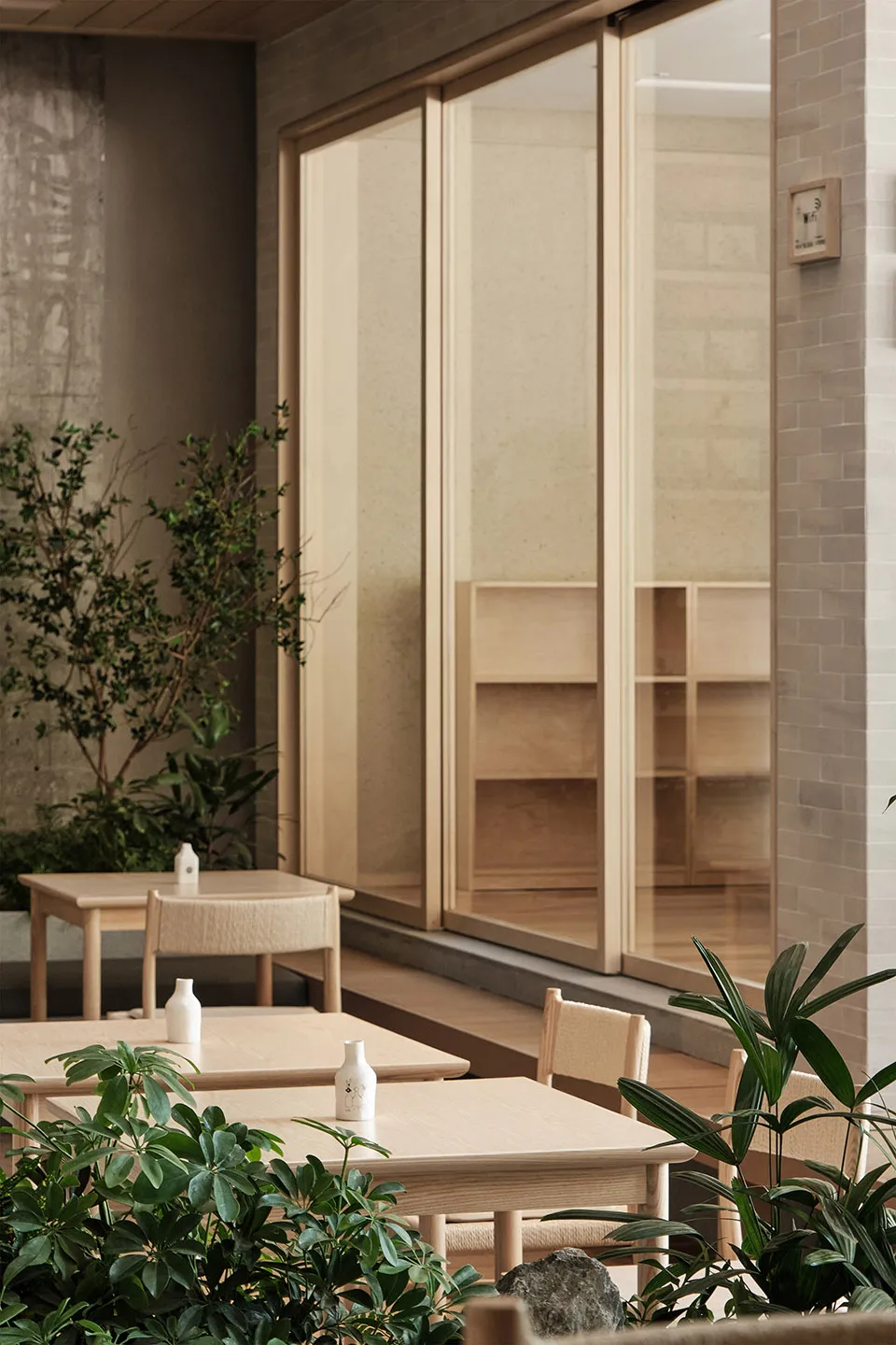 A minimalist restaurant interior with wood and glass materials and green plants.