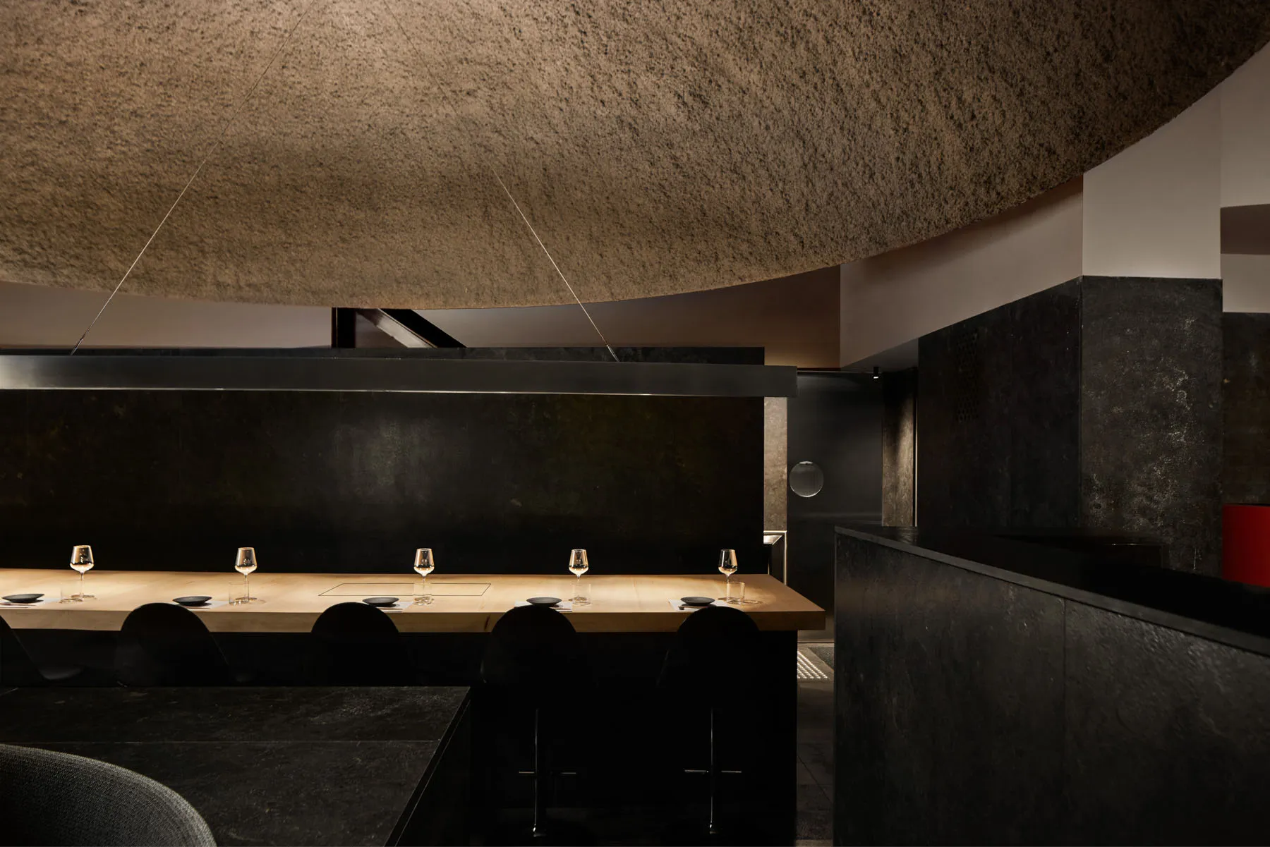 A minimalist restaurant interior with a long wooden table topped with wine glasses and plates, surrounded by black chairs and