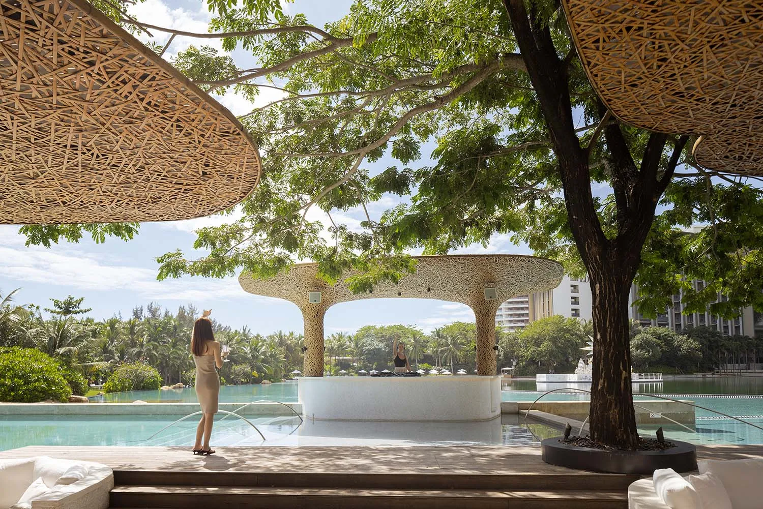 A modern poolside bar with bamboo architecture and a DJ playing music under a large tree.