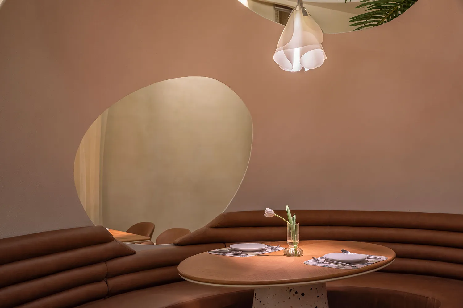 A modern restaurant interior with a curved leather bench seat and a table featuring a vase with a pink flower and cutlery.