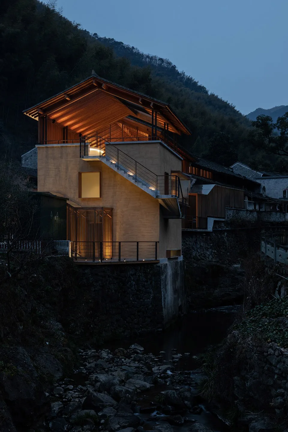 A modern residential house with concrete and wooden elements at dusk with mountains in the background.