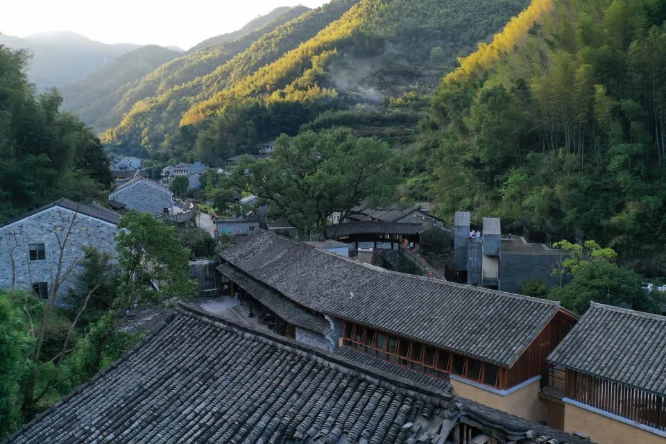 A traditional village with stone buildings and thatched roofs surrounded by lush greenery and mountains in the background.