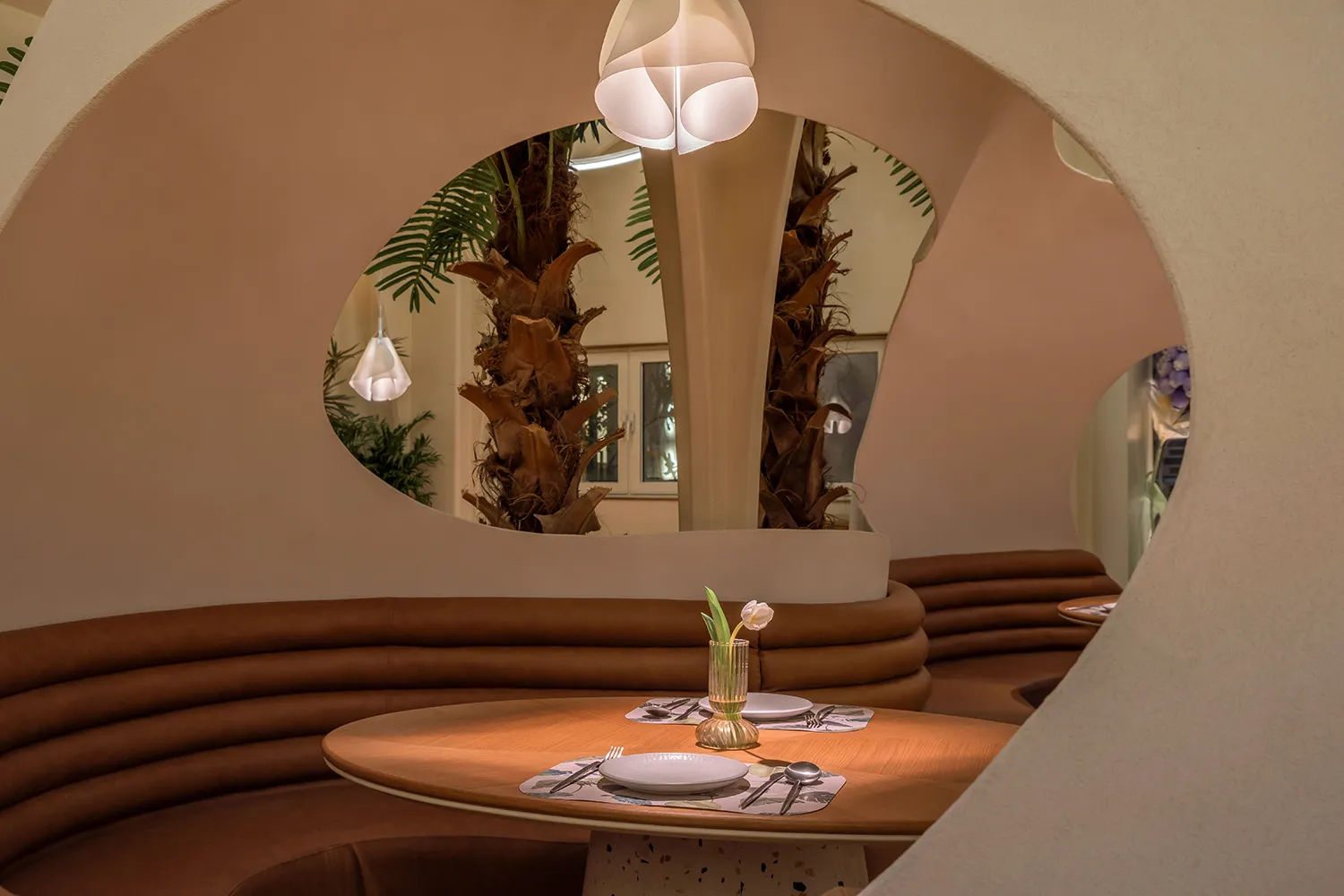 A modern restaurant setting featuring a round table with white plates, silverware, and a single white tulip in a vase, surrou