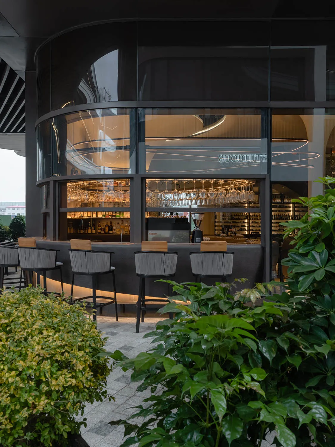 A modern restaurant bar featuring a glass partition, concrete furniture, and wine glasses hanging on the bar wall.