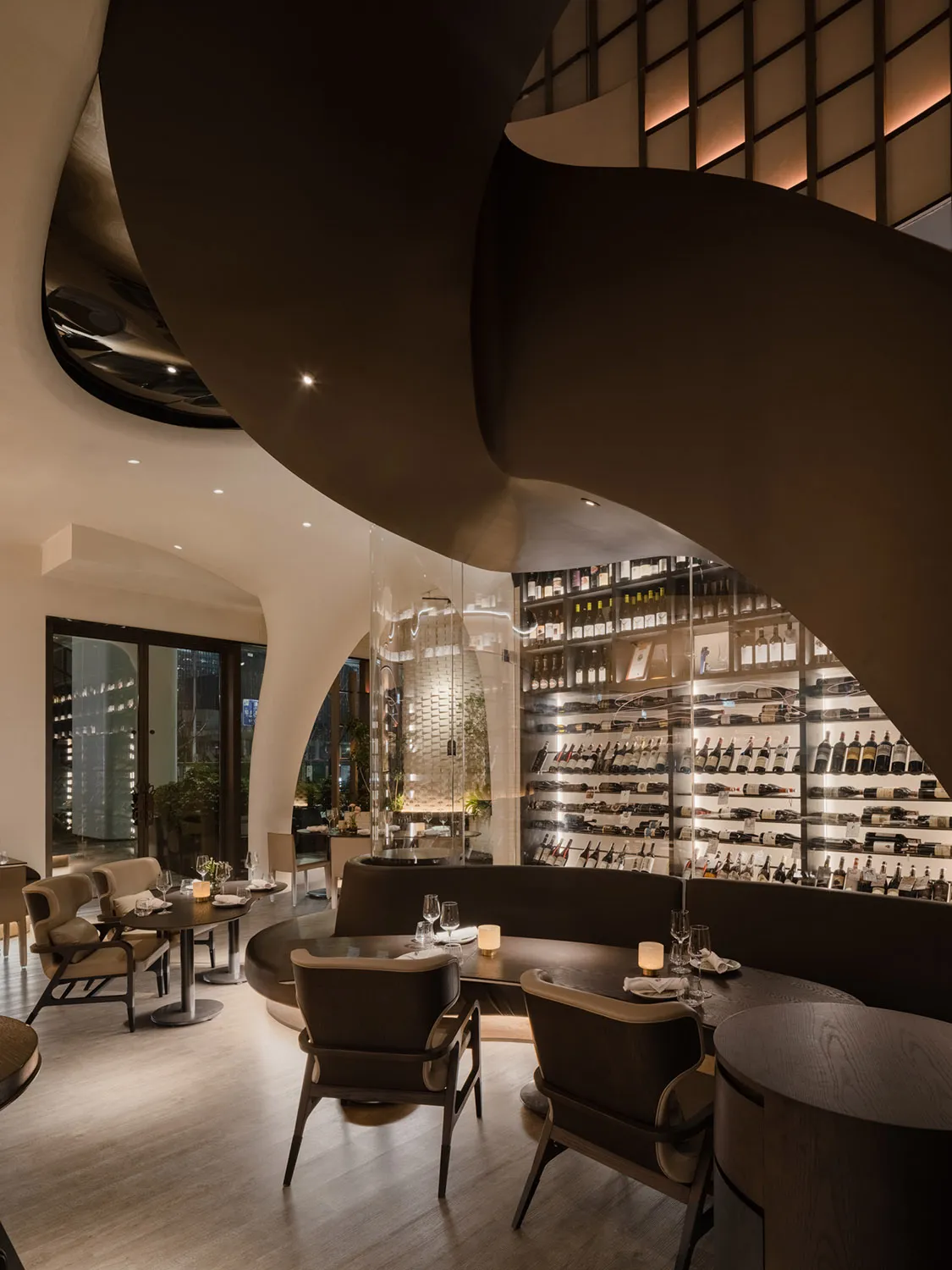 A modern restaurant interior with dark wood tables, glass wine shelves, and natural light through large windows.