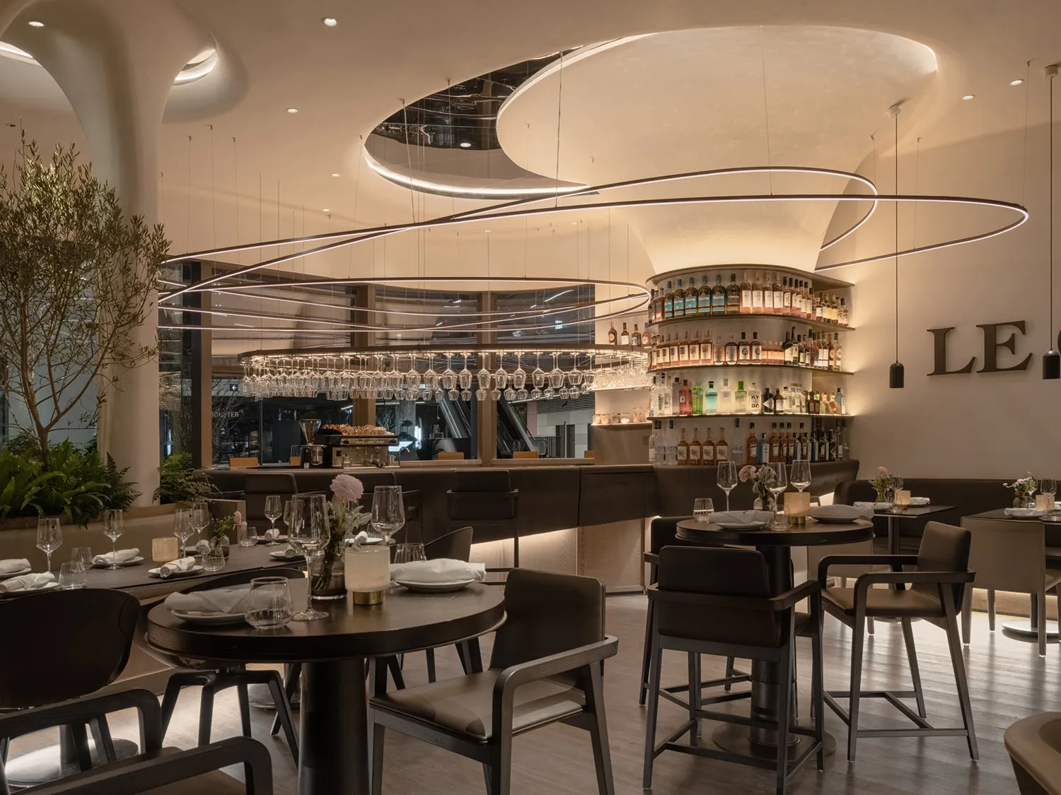 A modern restaurant interior featuring dark wood tables, leather chairs, glass shelves, and floral arrangements.
