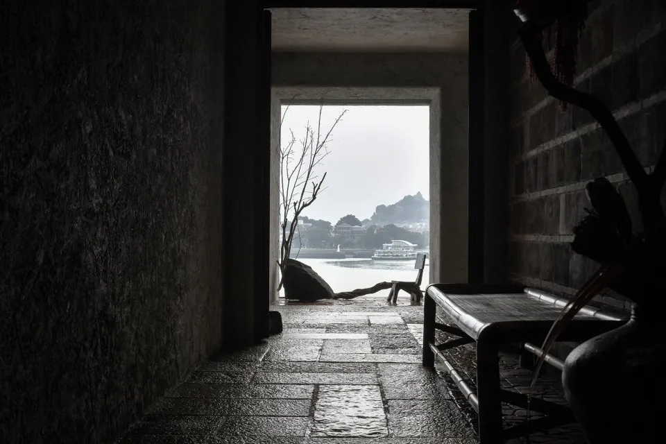 A dark, stone-walled interior with a wooden bench and view of a calm river and distant buildings.