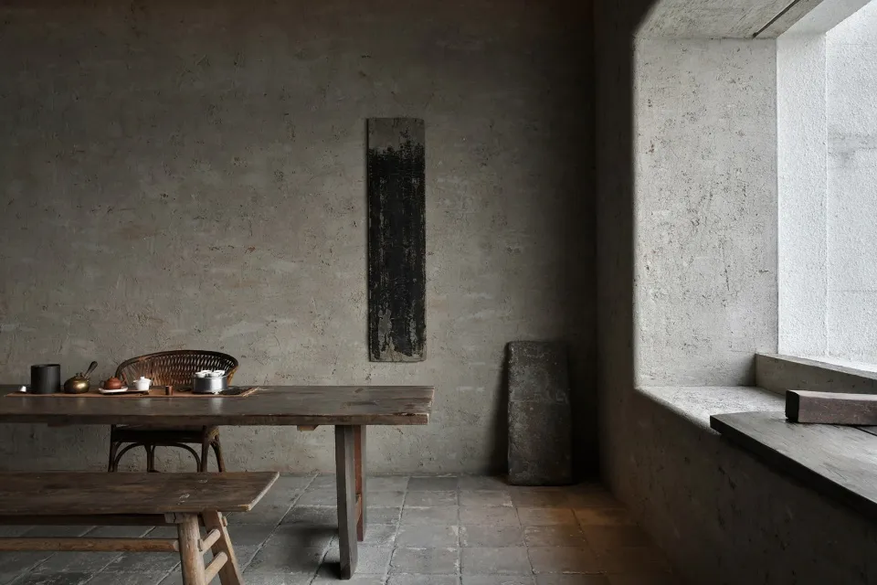 A minimalist kitchen features a wooden table, a concrete wall, and natural light from a window.