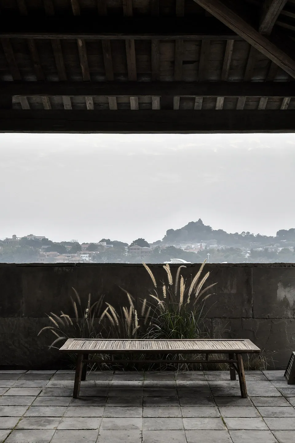 A wooden bench sits on a tiled rooftop with a view of a distant cityscape and a hill.