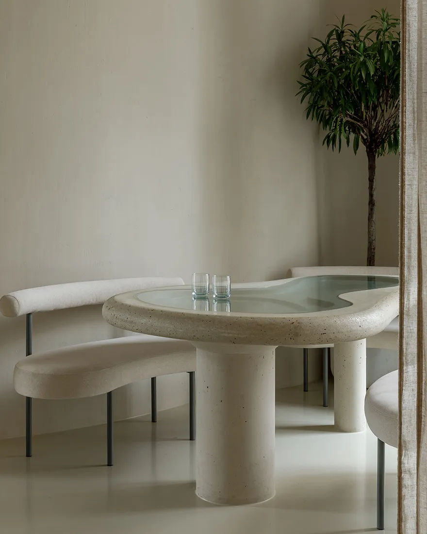 Modern minimalist dining area with a round, curved concrete table and matching benches, featuring a sleek, glass-top centerpi