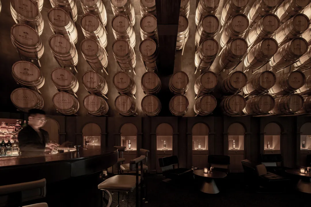 A dimly lit, industrial-style bar interior featuring numerous stacked barrels on the walls, creating a unique and textured ap