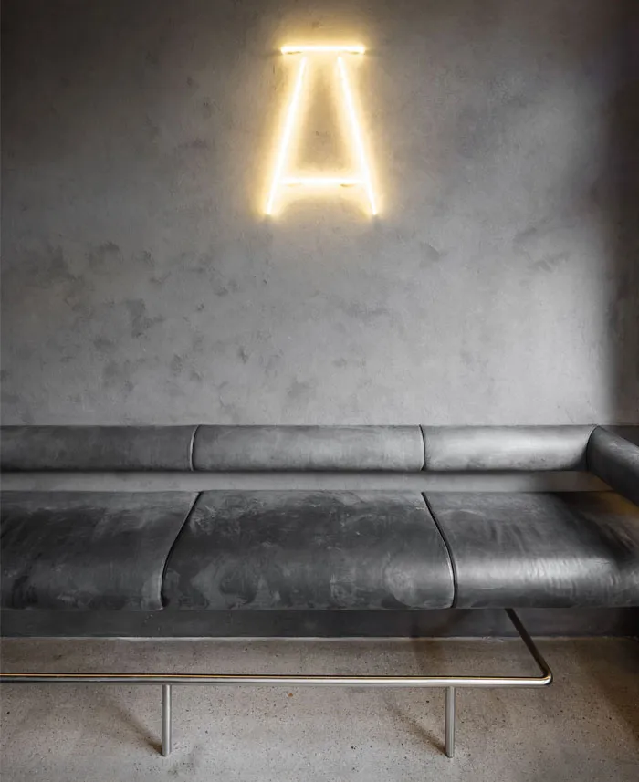 Industrial-style interior with concrete walls, metal seating, and a neon letter 'A' light fixture.