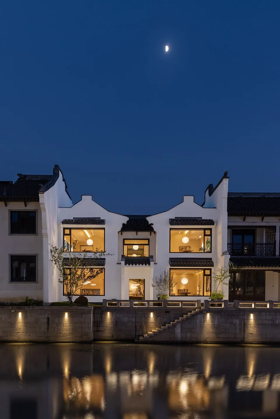 Modern building with black roof tiles and white-painted exterior reflected in water at dusk with illuminated windows.