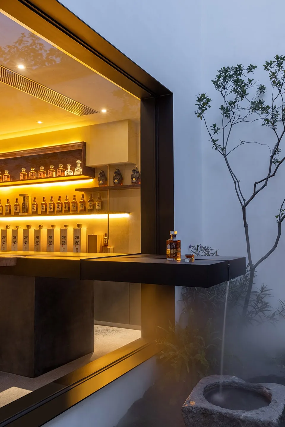 A sleek, modern bar counter with a concrete base and metal structure, featuring illuminated shelves and a water feature in th