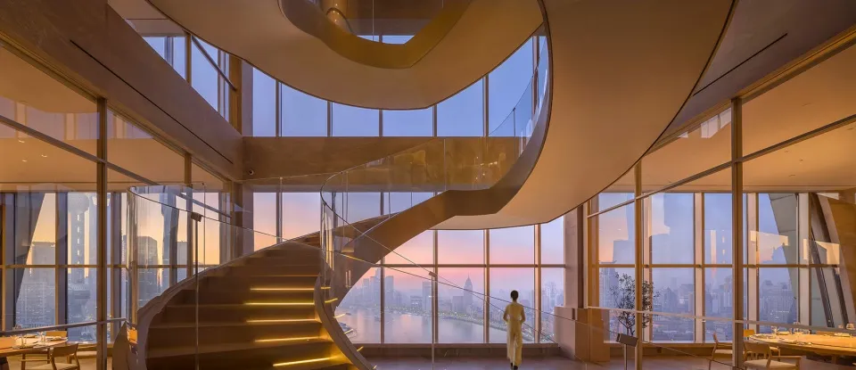 A glass-enclosed space viewed from within, featuring a spiral staircase and a panoramic cityscape.