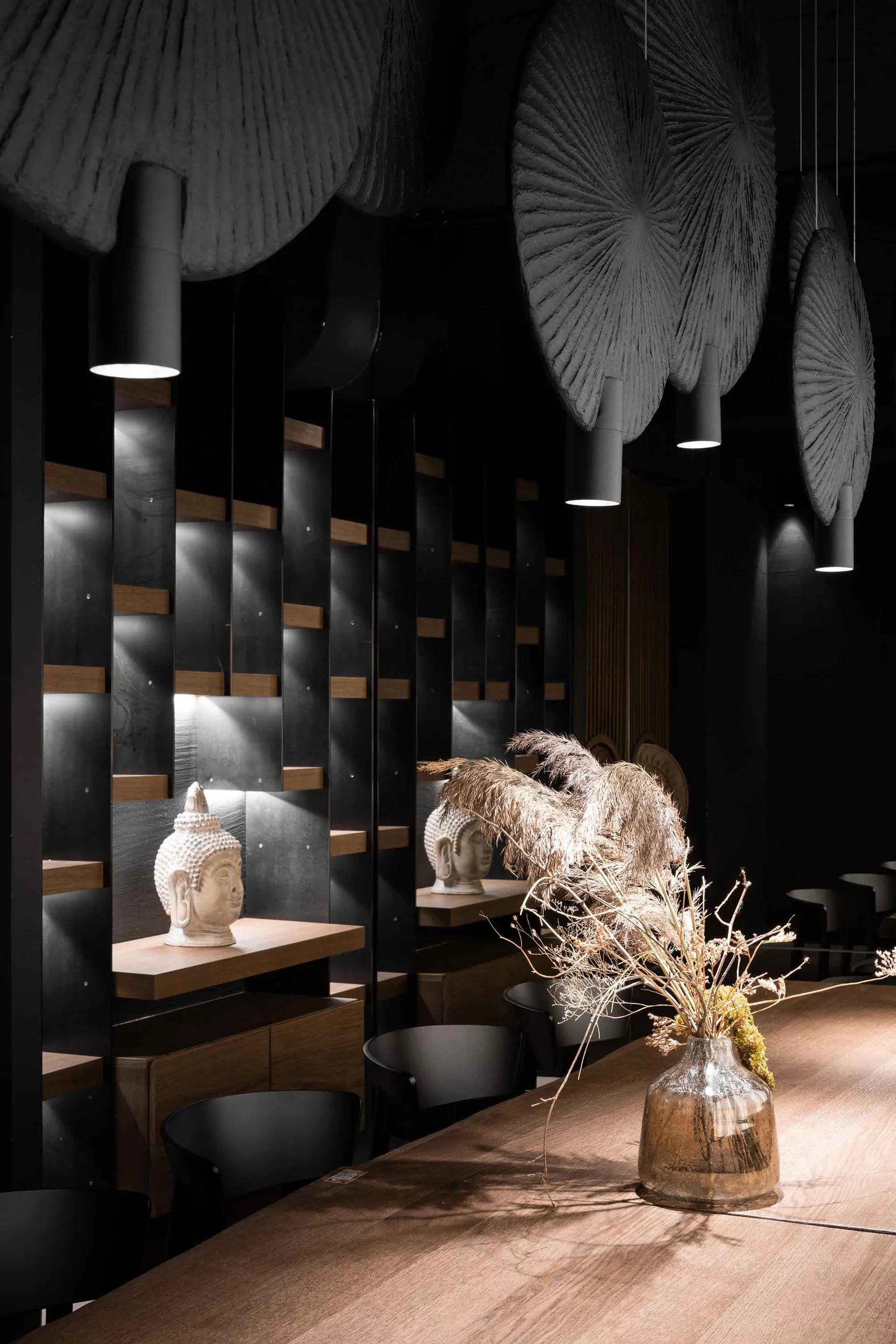 A modern restaurant interior with wooden accents and minimalist design, featuring Buddha statues and dried plants.