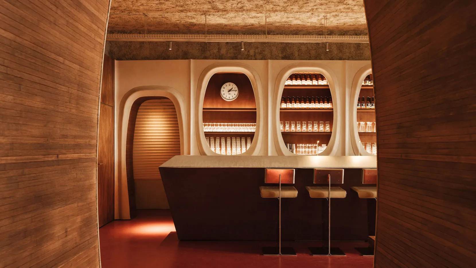 A modern bar interior with a wooden counter, arched walls, and illuminated shelves.