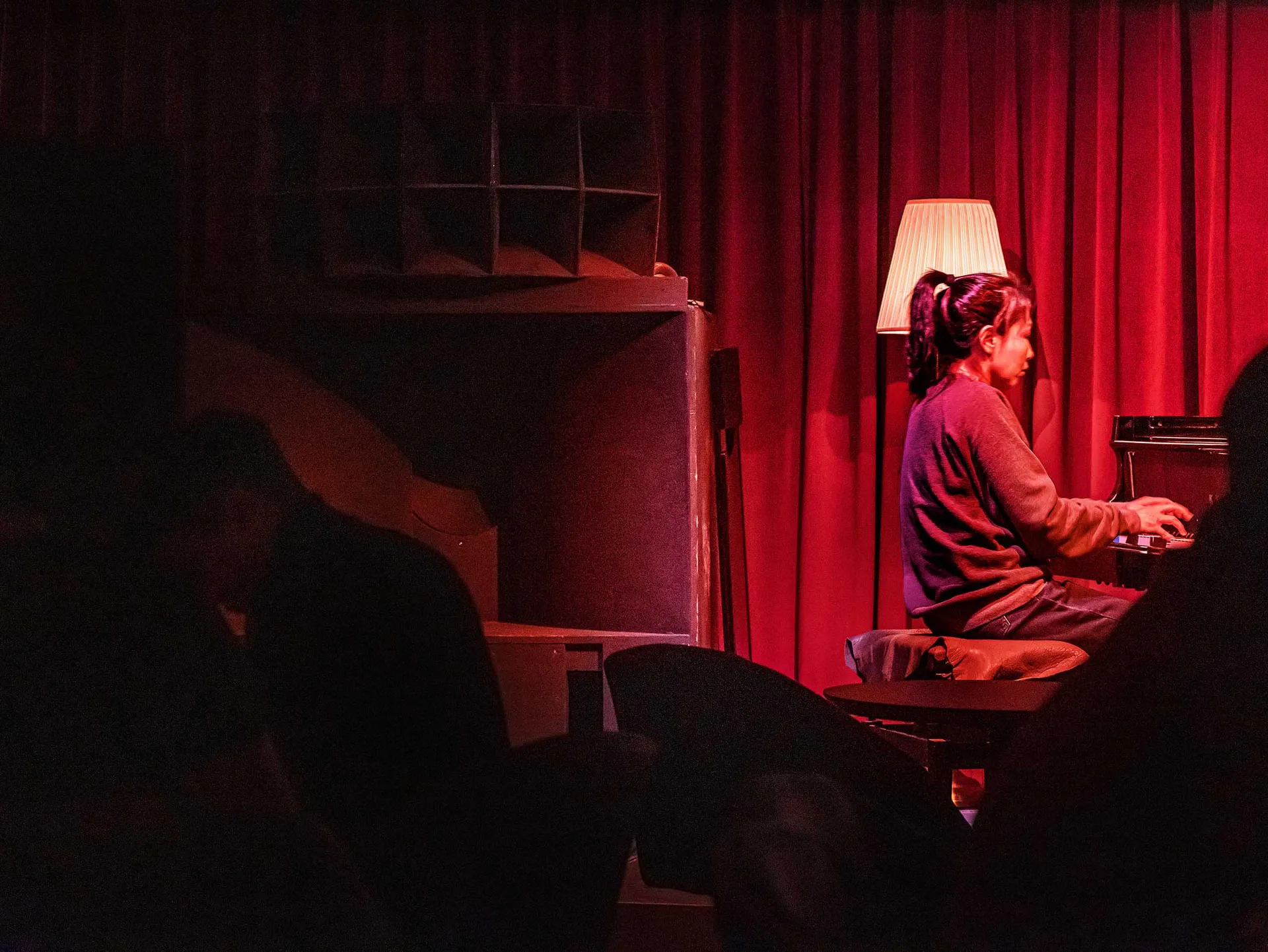 A woman playing the piano under a red curtain with a lamp illuminating the scene.