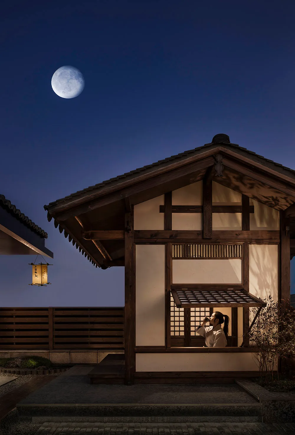 A traditional Japanese wooden structure with a paper lantern hanging outside, under a clear night sky with a full moon.