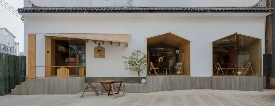 A minimalist façade with wooden accents and glass panels of a restaurant with a concrete base and a tree in front of it.