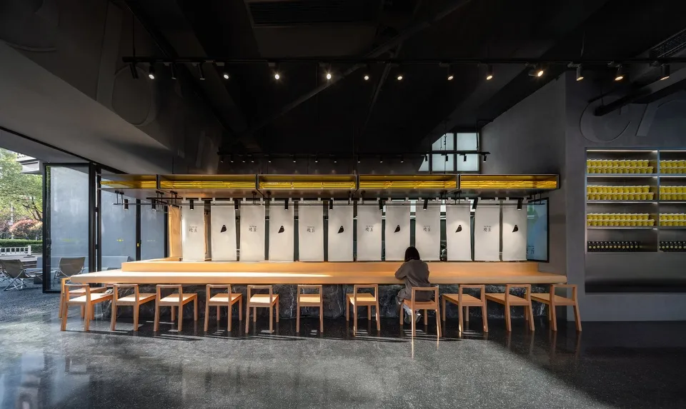A modern restaurant interior featuring a long wooden counter with seating, minimalist black decor, and illuminated shelves.