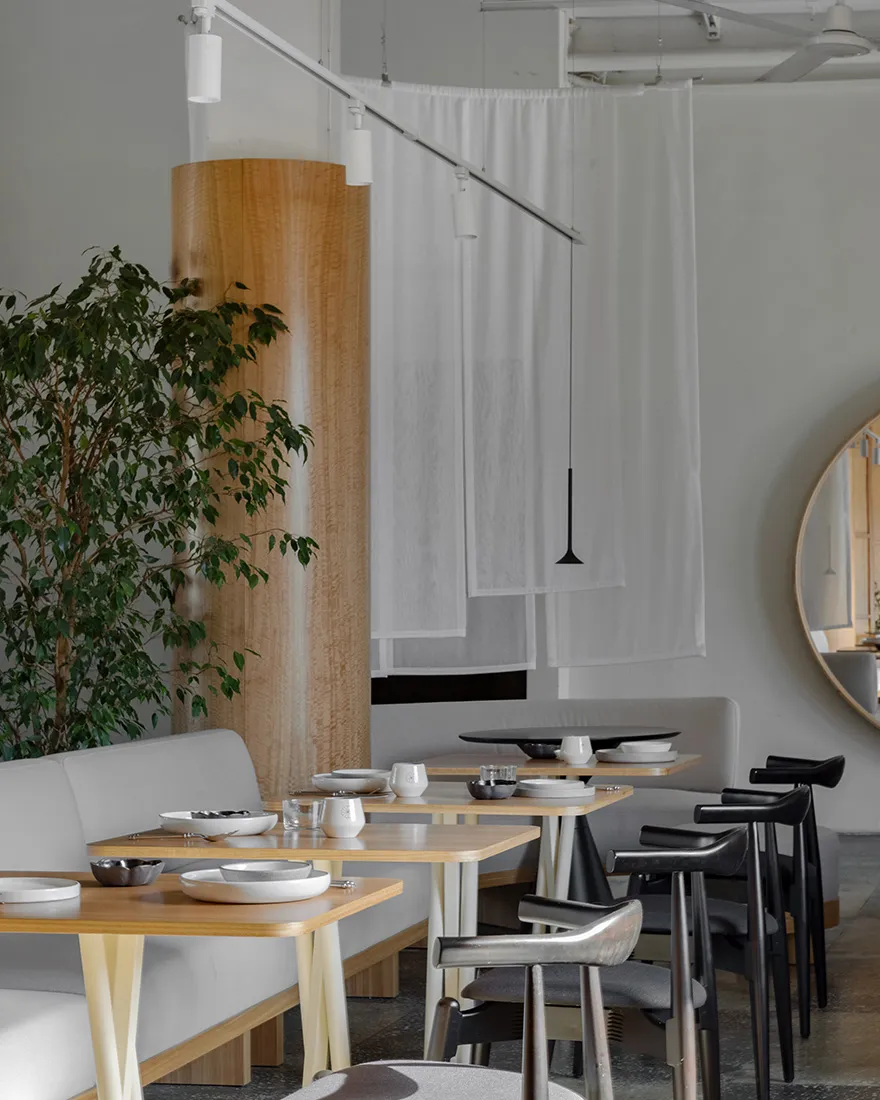 A modern restaurant interior with minimalist design features, including wooden tables, fabric seating, and metallic accents.