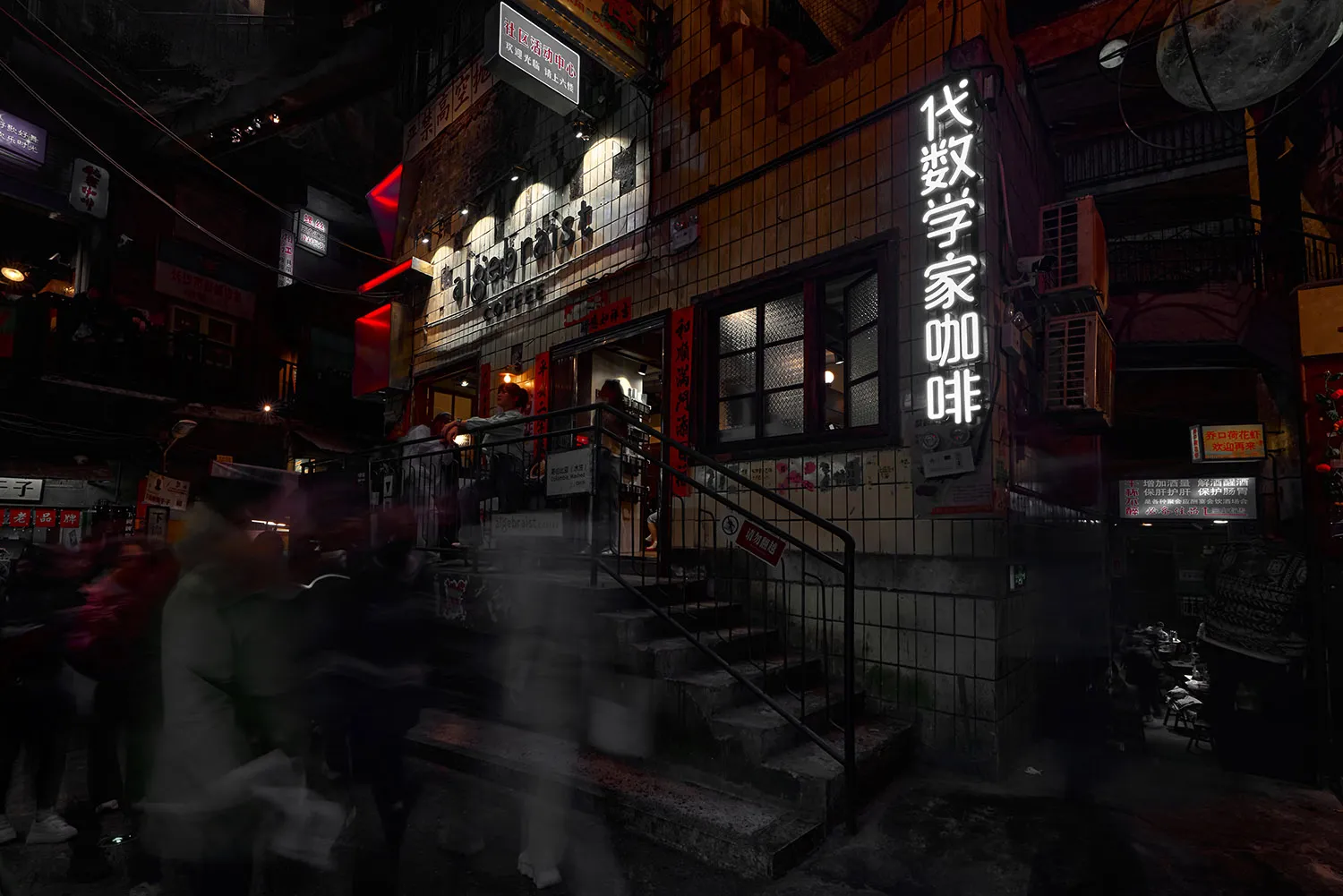 A bustling urban street at night, featuring a tiled storefront with illuminated signage and a metal staircase.