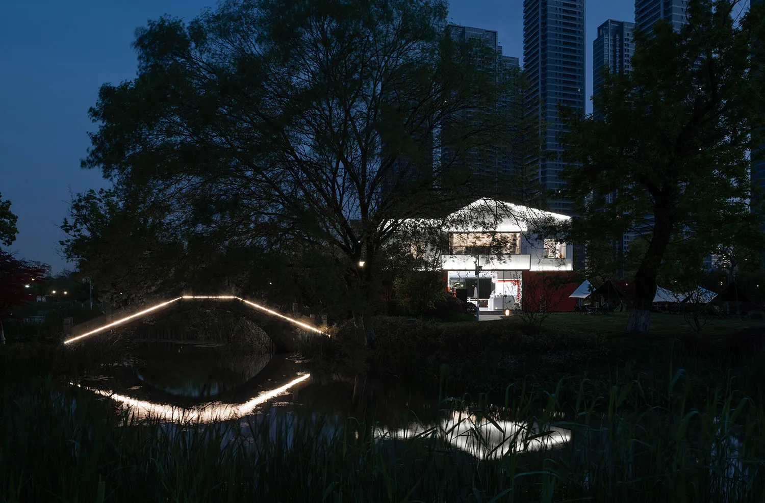 A modern house at night with illuminated lights and a pond with a bridge.