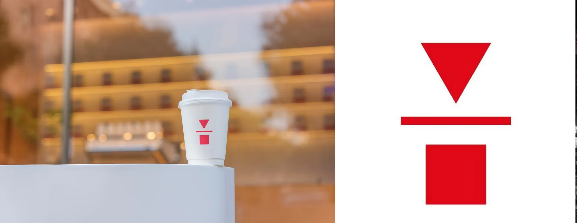 A white paper coffee cup with a red triangle and square logo sits on a white table in a modern coffee shop.