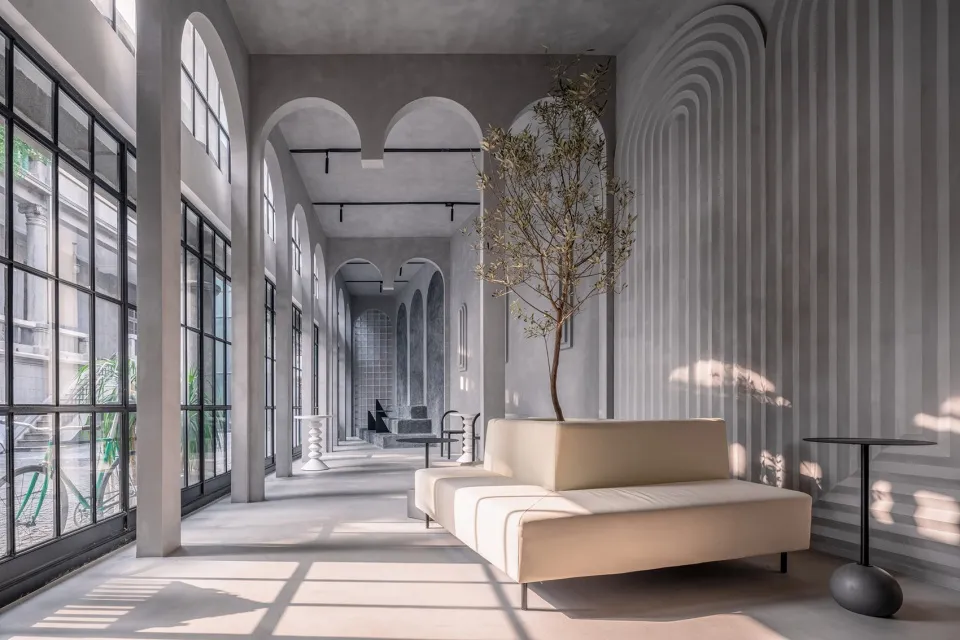 A minimalist lobby with concrete walls and floor, featuring an olive tree in a white pot and a sleek, modular seating area.