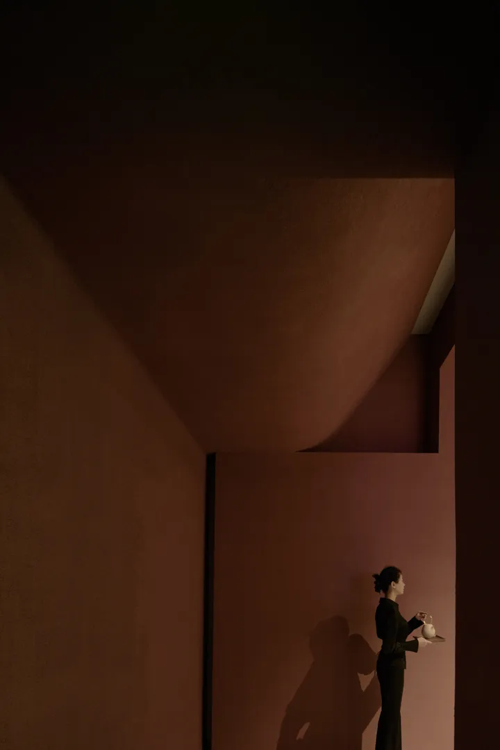A minimalist interior space featuring exposed concrete walls and a single figure holding a teapot on a tray.