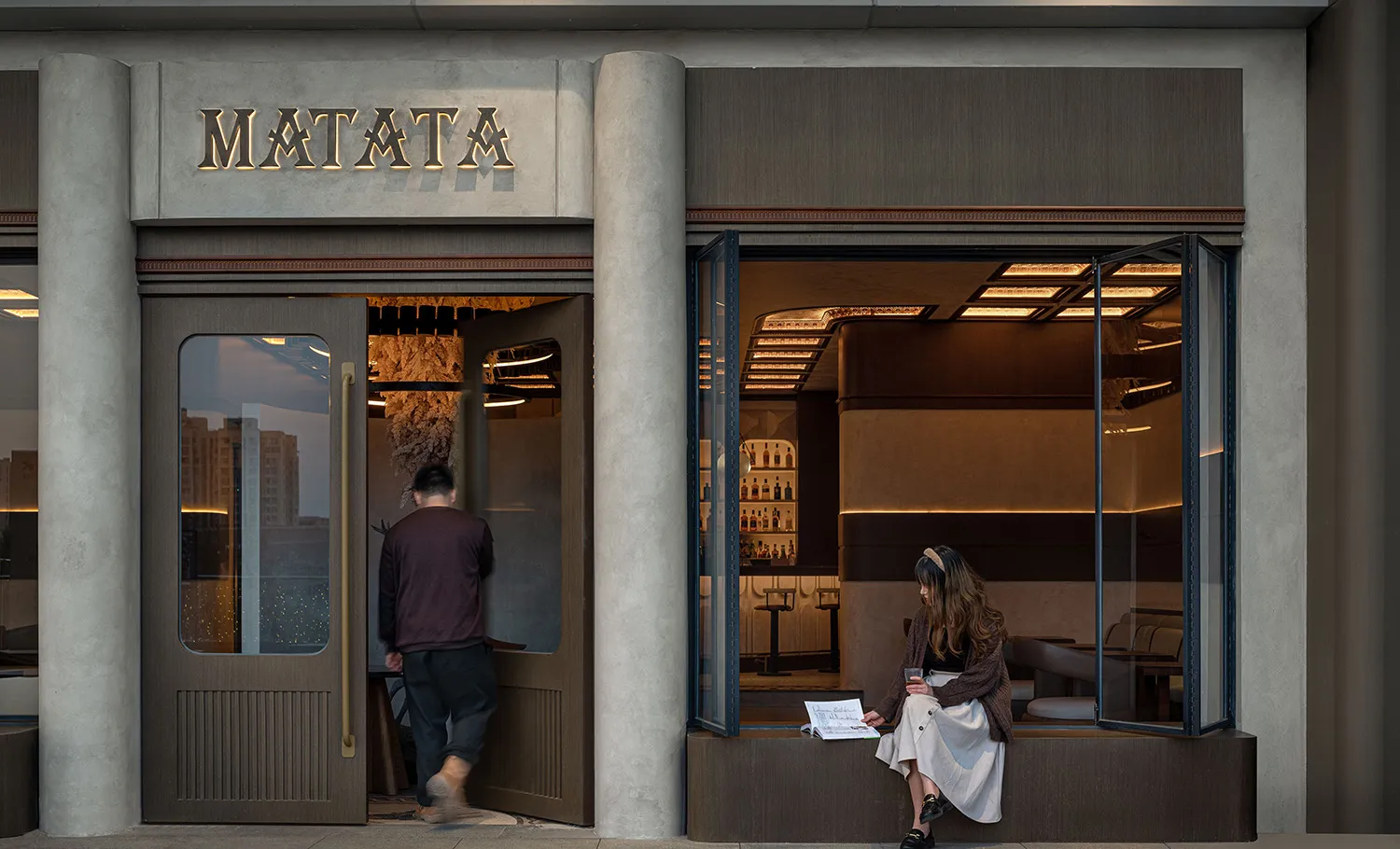 A casual-dining restaurant with a concrete exterior, featuring a person sitting in a window seat and another entering the est