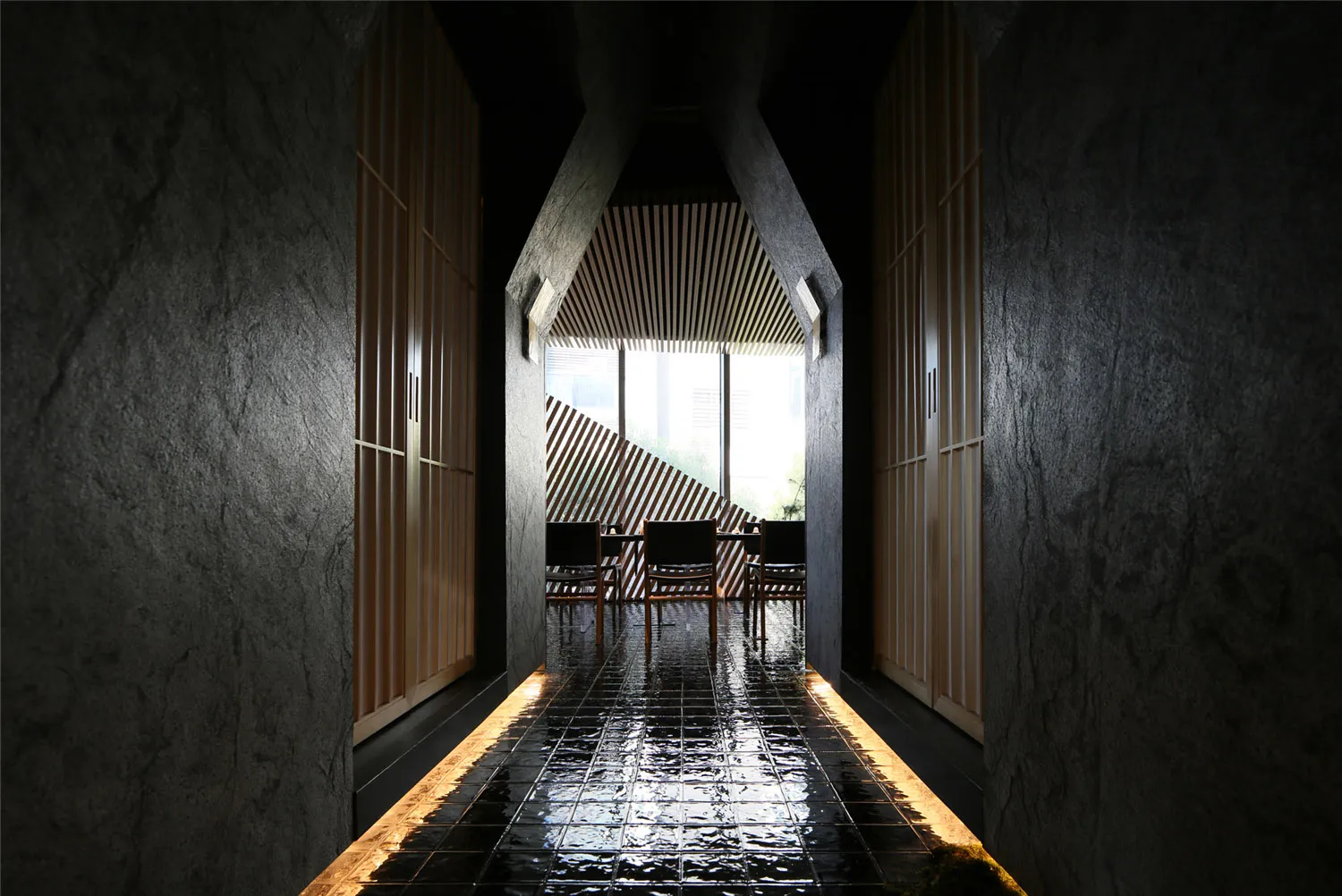 A minimalist waiting area with concrete walls, wood furniture, and a view through a tall window.