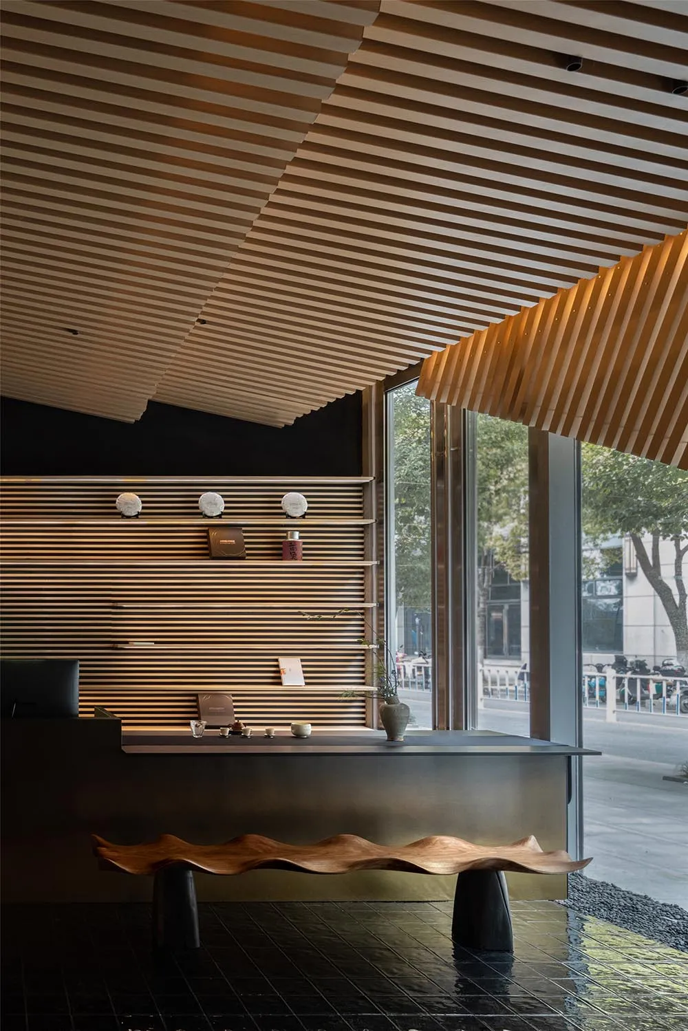 A modern reception desk made of wood with a wavy design, featuring a sleek black countertop and a curved wooden bench under t