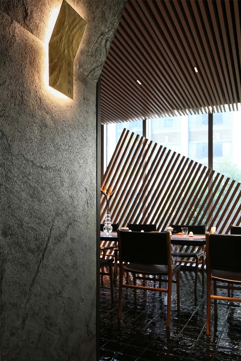 A modern restaurant interior featuring a wooden wall and ceiling paneling with a vase on a dining table and a window with str