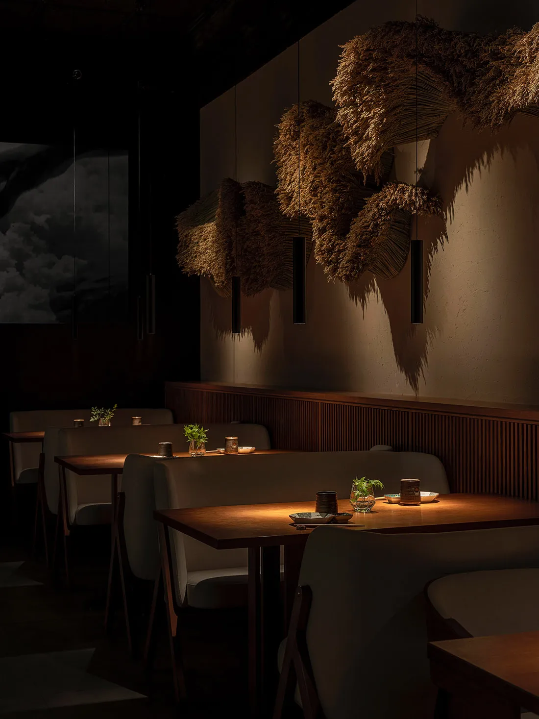 Modern minimalist dining room with wood tables, upholstered benches, fabric and dried-grass accents, and dim lighting.