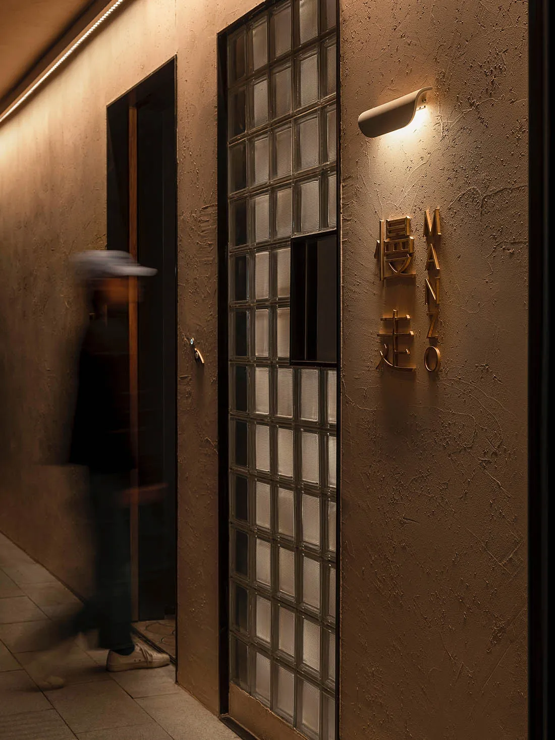 A modern restaurant entryway featuring glass block walls and a brass nameplate mounted on a textured beige wall.