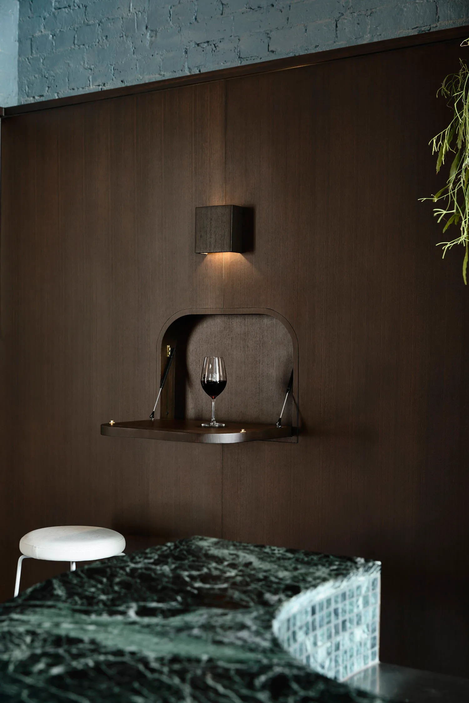 A minimalist wine bar designed for a small space, featuring a wooden shelf with a glass of red wine illuminated by a wall-mou