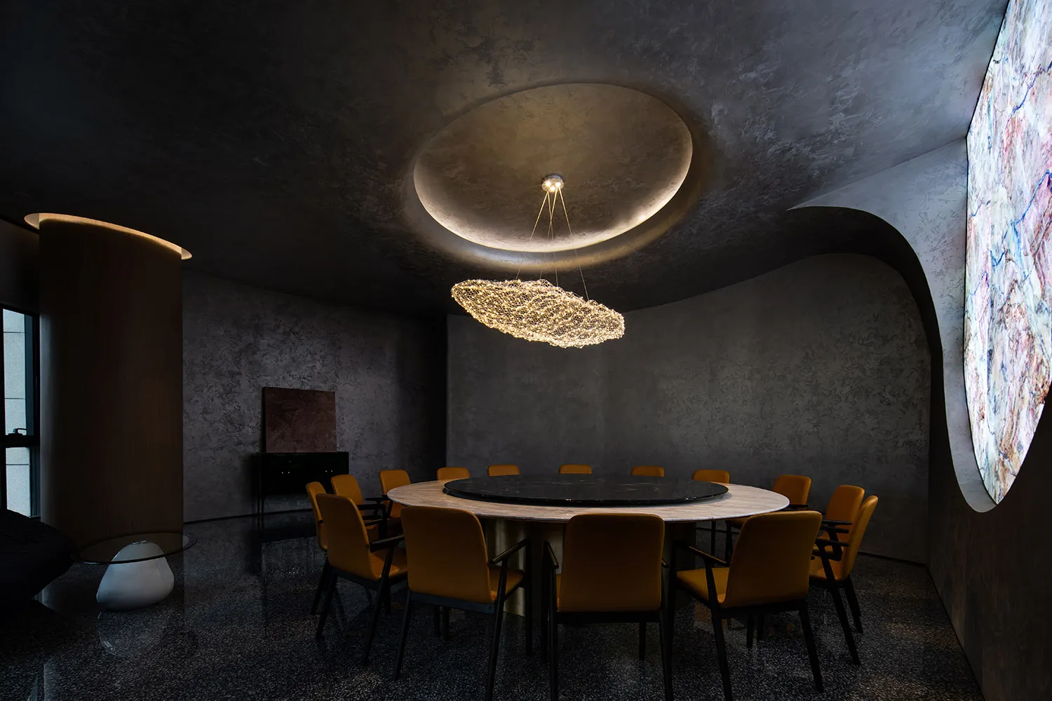 Modern conference room featuring a round table surrounded by chairs, with a chandelier, marble floor, and concrete walls.
