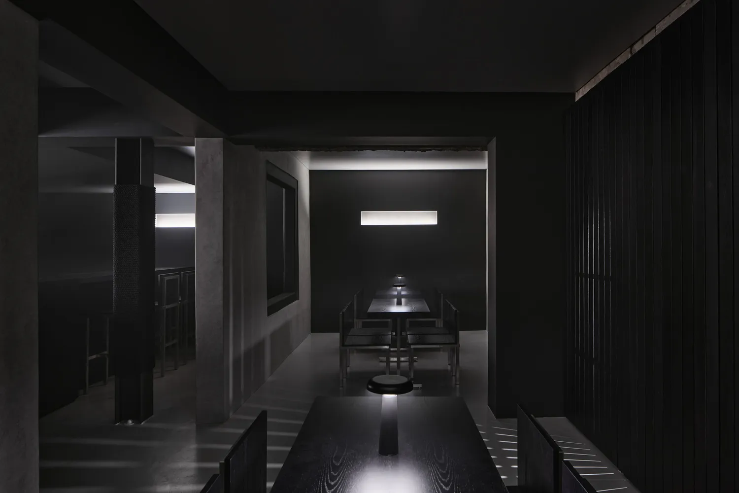 A minimalist restaurant interior with black walls and tables, illuminated by a single light source.