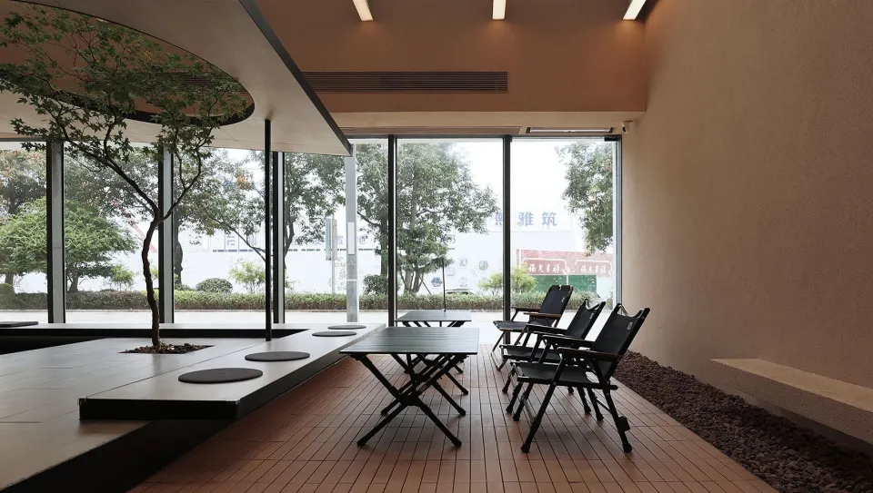 Modern restaurant interior with a wooden floor, tables, and chairs, featuring large windows with trees and greenery visible o