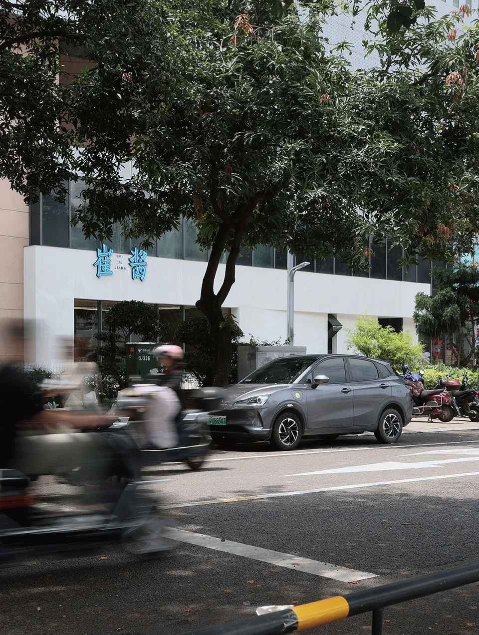現代都市景觀，灰色汽車停在樹下，背景有其他車輛與建築物。