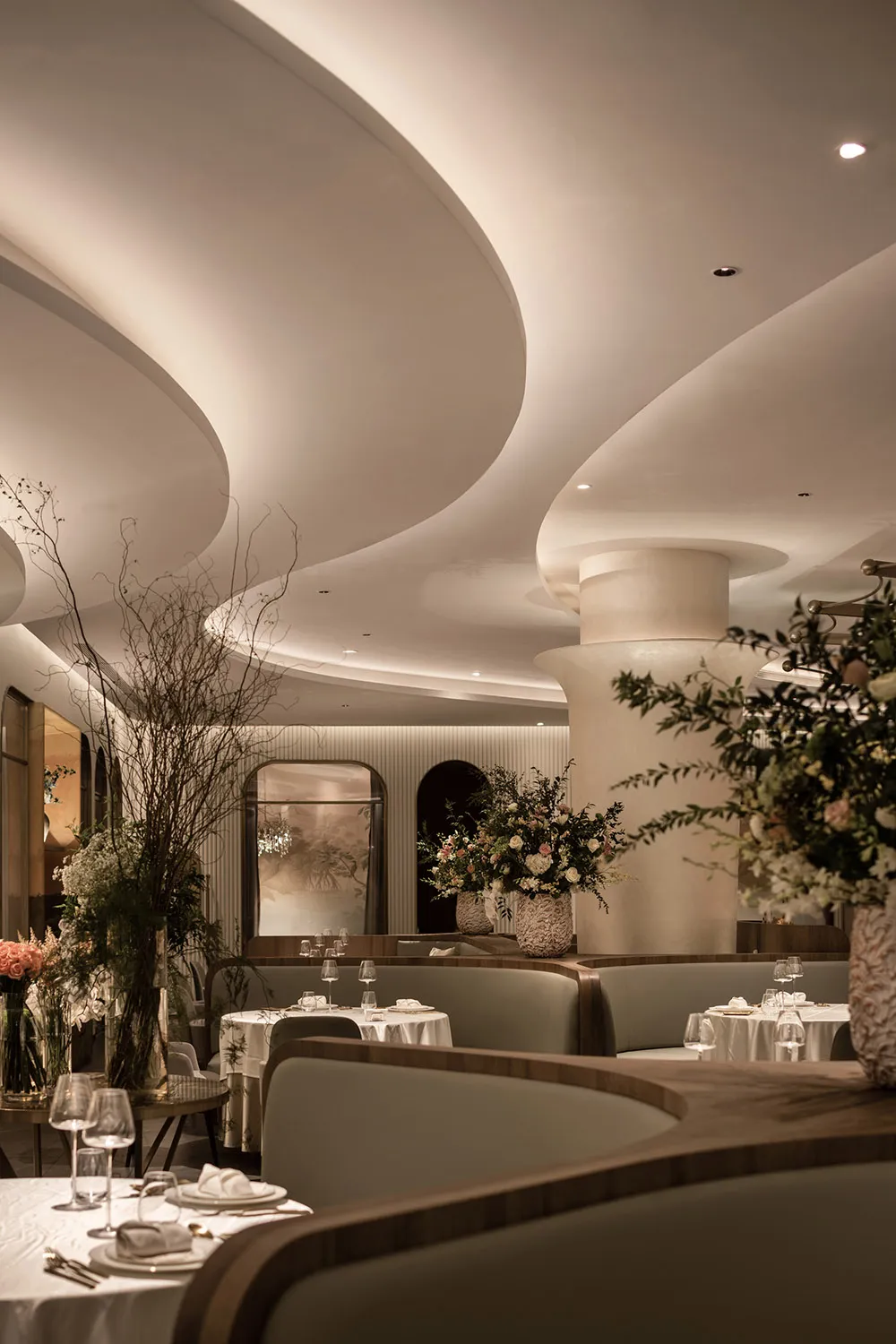 Modern restaurant dining room with white and gray decor, curved ceiling, and floral arrangements.
