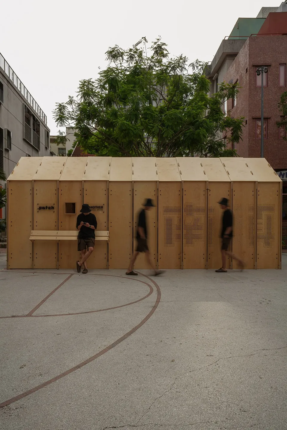 A minimalist wooden shelter in an urban setting, with people walking past.